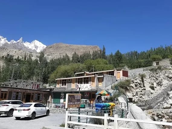 A roadside building and forecourt below a wooded slope, with snow peaks at the left