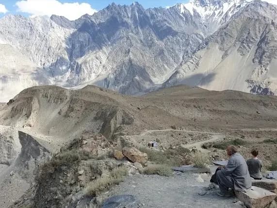 Two people sitting on a rock above a trail, looking out at the Karakoram peaks