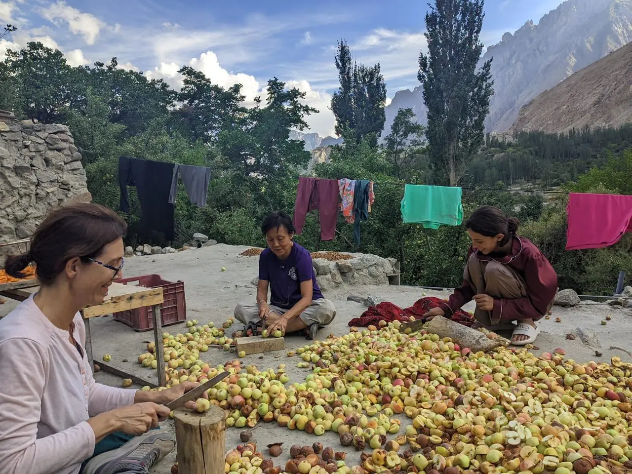 Three people sitting on a roof terrace cutting apricots to dry, with laundry lines and the mountains behind