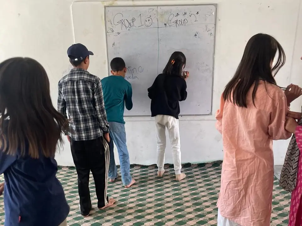 Children writing at a whiteboard split into Group 1 and Group 2, with classmates watching from the mat